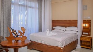 Habitación de hotel con cama de madera, ropa de cama blanca, mesita de noche de madera con una botella y posavasos, y una lámpara fijada a la pared.