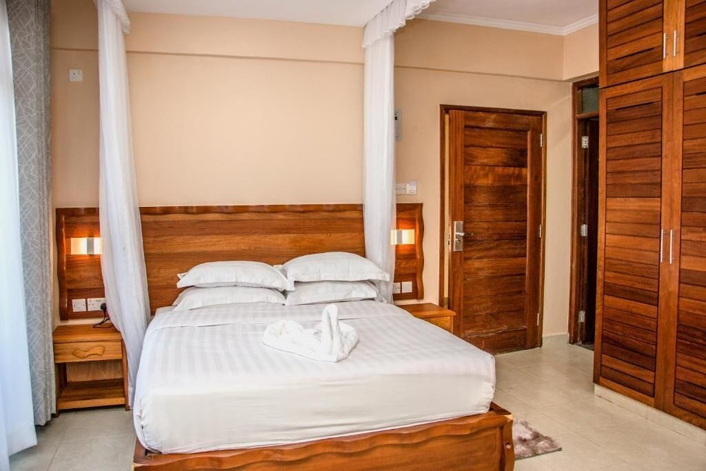 Una cama bien tendida con un almohadón con forma de cisne blanco, cabecero de madera y lámparas de noche.