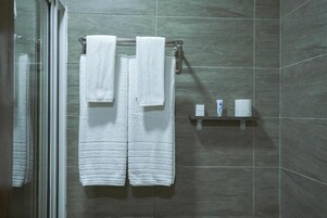 Un baño con mampara de vidrio, dos toallas blancas colgadas en un perchero y un pequeño estante con artículos de aseo.