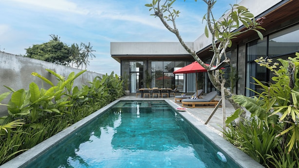 Uma área moderna de piscina ao ar livre, com espreguiçadeira e mesa, rodeada por vegetação exuberante e um céu azul límpido.