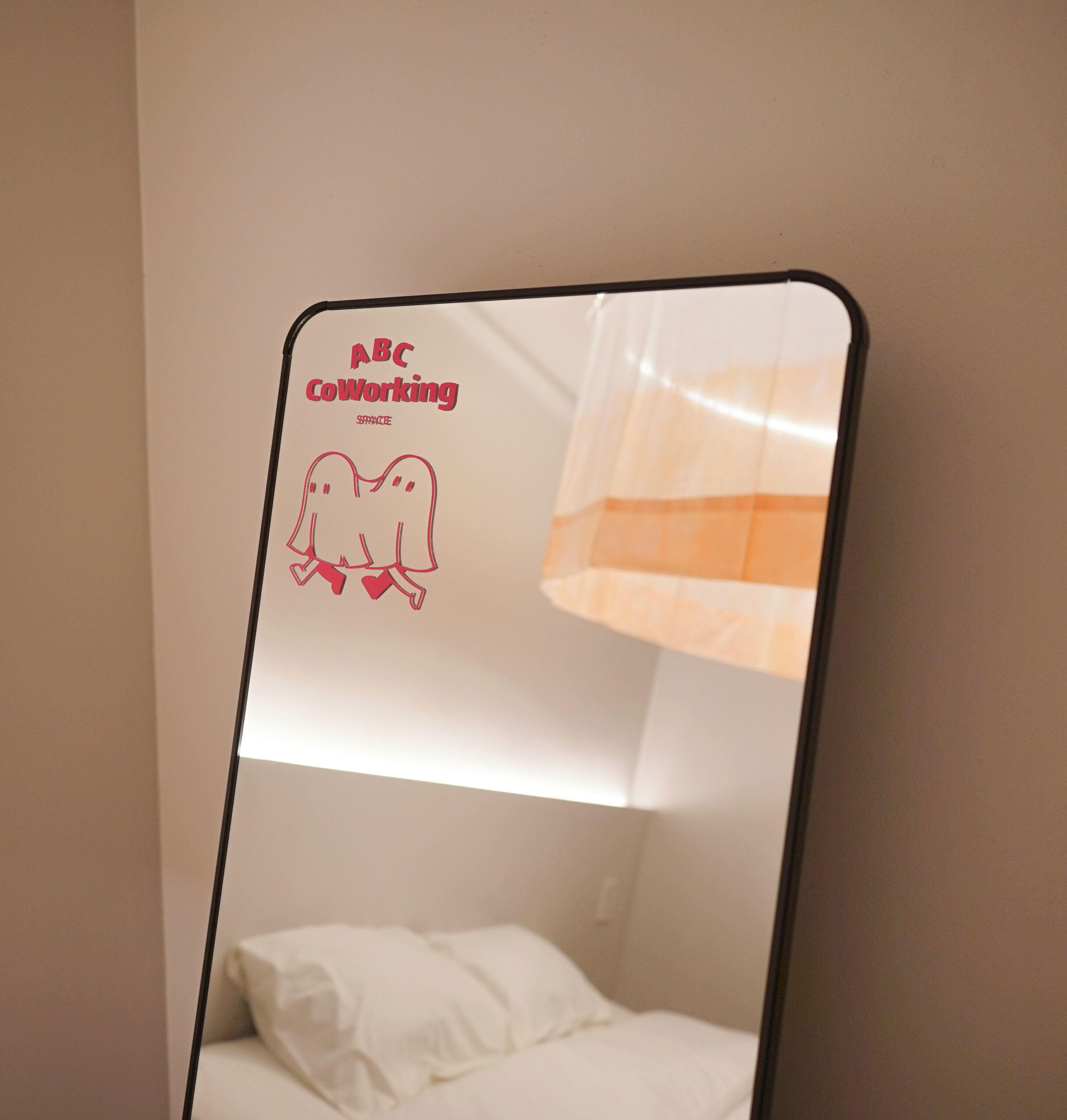 A room with a bed, a wall-mounted mirror, and a sign with 'ABC Coworking SPACE' and a drawing of two figures holding hands.
