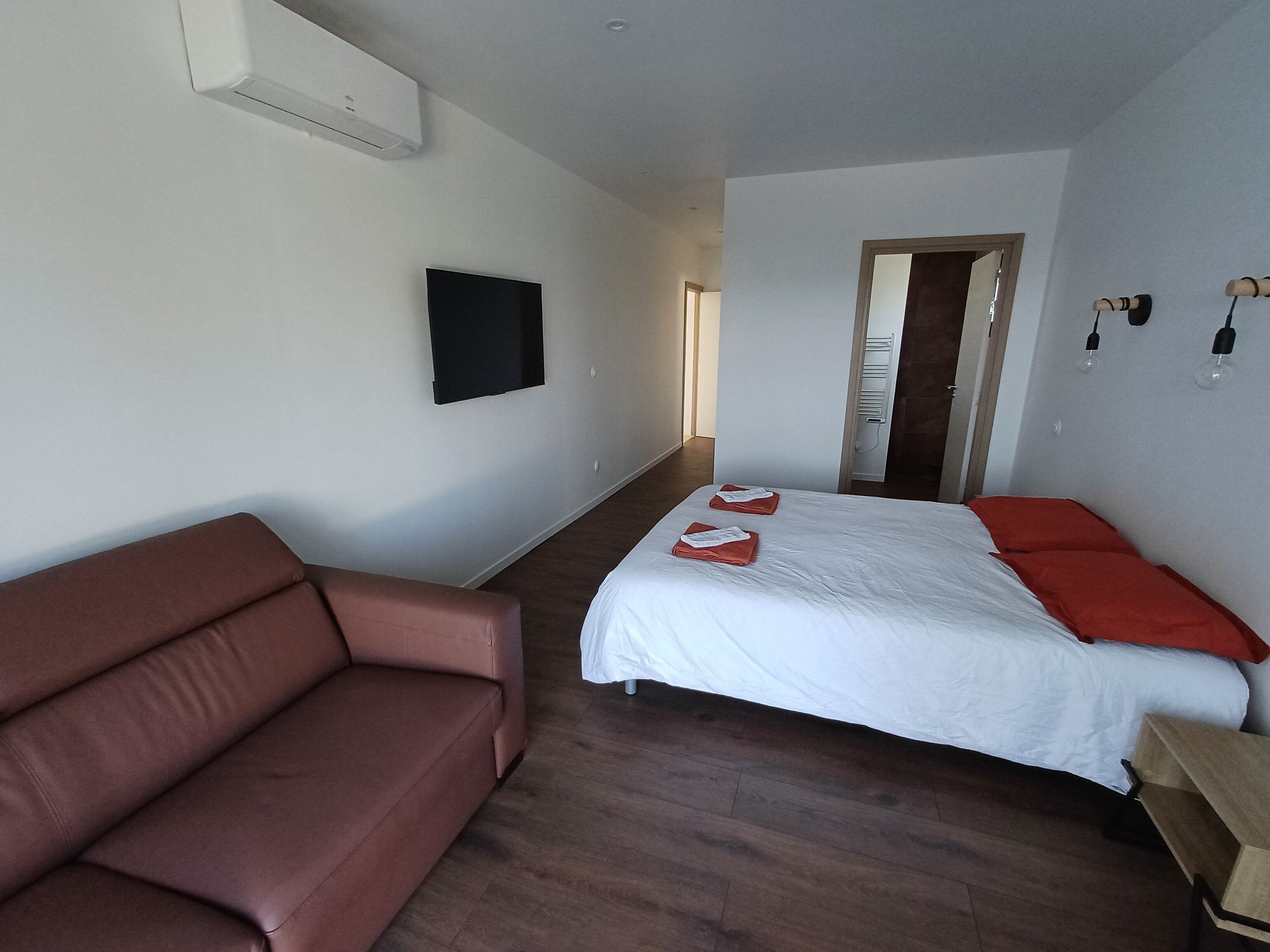 Une chambre d’hôtel avec un canapé marron, un lit avec une literie blanche et rouge, un téléviseur à écran plat fixé au mur et un plancher en bois.