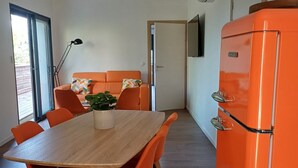 Ein Essbereich mit einem Holztisch und orangefarbenen Stühlen, einer Couch mit einem bunten Kissen und einem leuchtend orangefarbenen Kühlschrank.