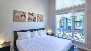 A bedroom with a large window, a bed with white bedding, and three wall-mounted artworks.
