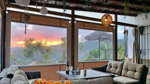 Una habitación con un ventanal grande que da a un paisaje montañoso al atardecer, un sofá, una mesa con una botella y copas, y plantas colgantes.