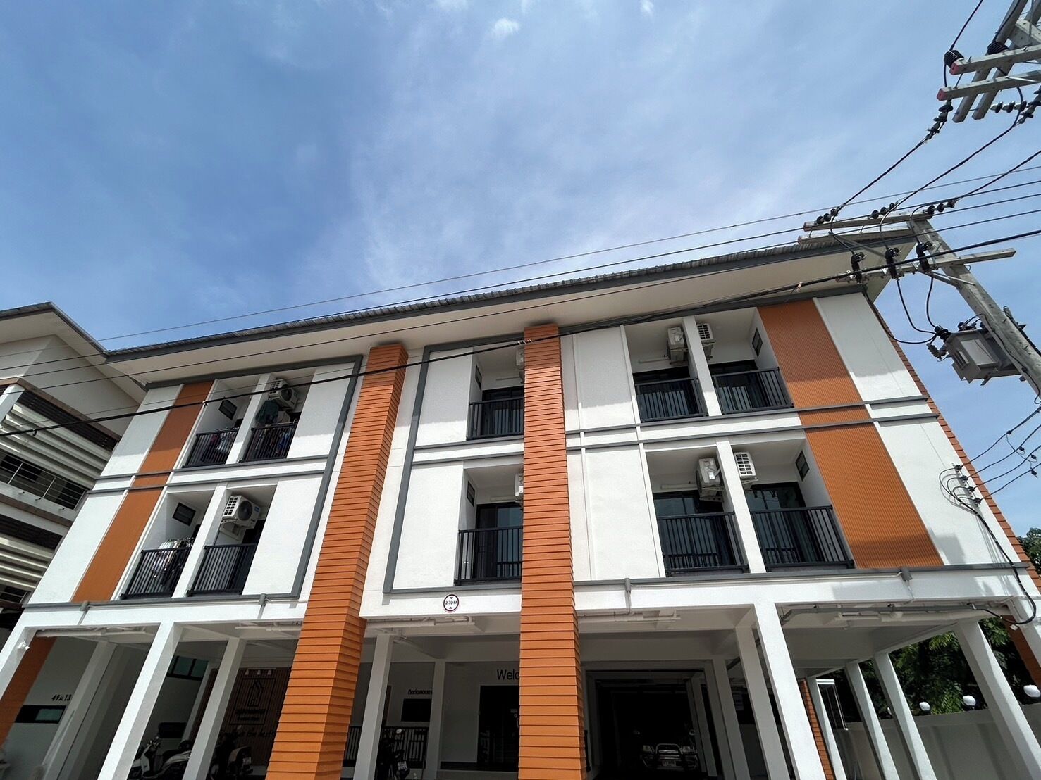 A multi-story building with a white facade and orange accents, featuring balconies and air conditioning units.