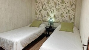 Quarto com duas camas de solteiro, cada uma com travesseiro verde, uma mesinha de cabeceira com abajur e papel de parede estampado na parede.