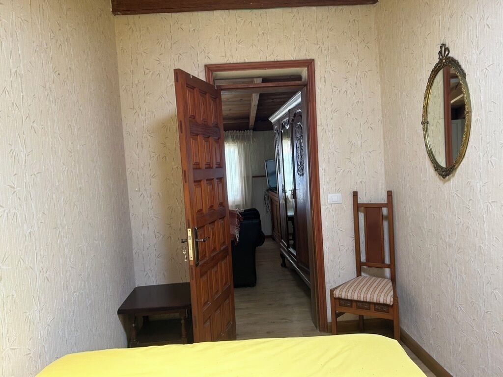 Quarto com porta de madeira, cama com colcha amarela, uma cadeira e um espelho na parede.