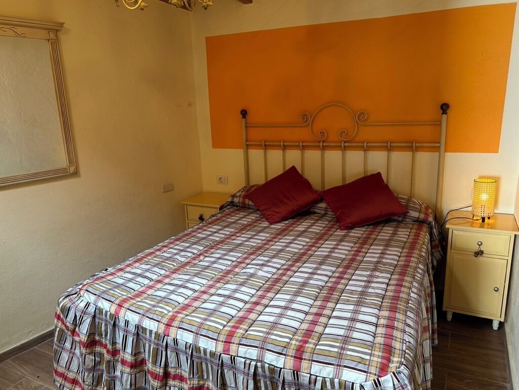 Een bed met een geruit sprei, twee rode kussens, een nachtkastje met een lamp, en een muur met een raam en een fotolijst.