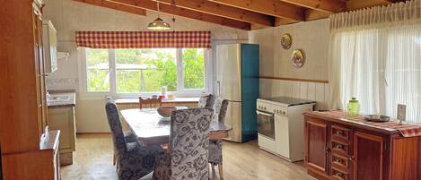 A kitchen with wooden cabinets, a dining table with chairs, a refrigerator, a stove, and a window with curtains.