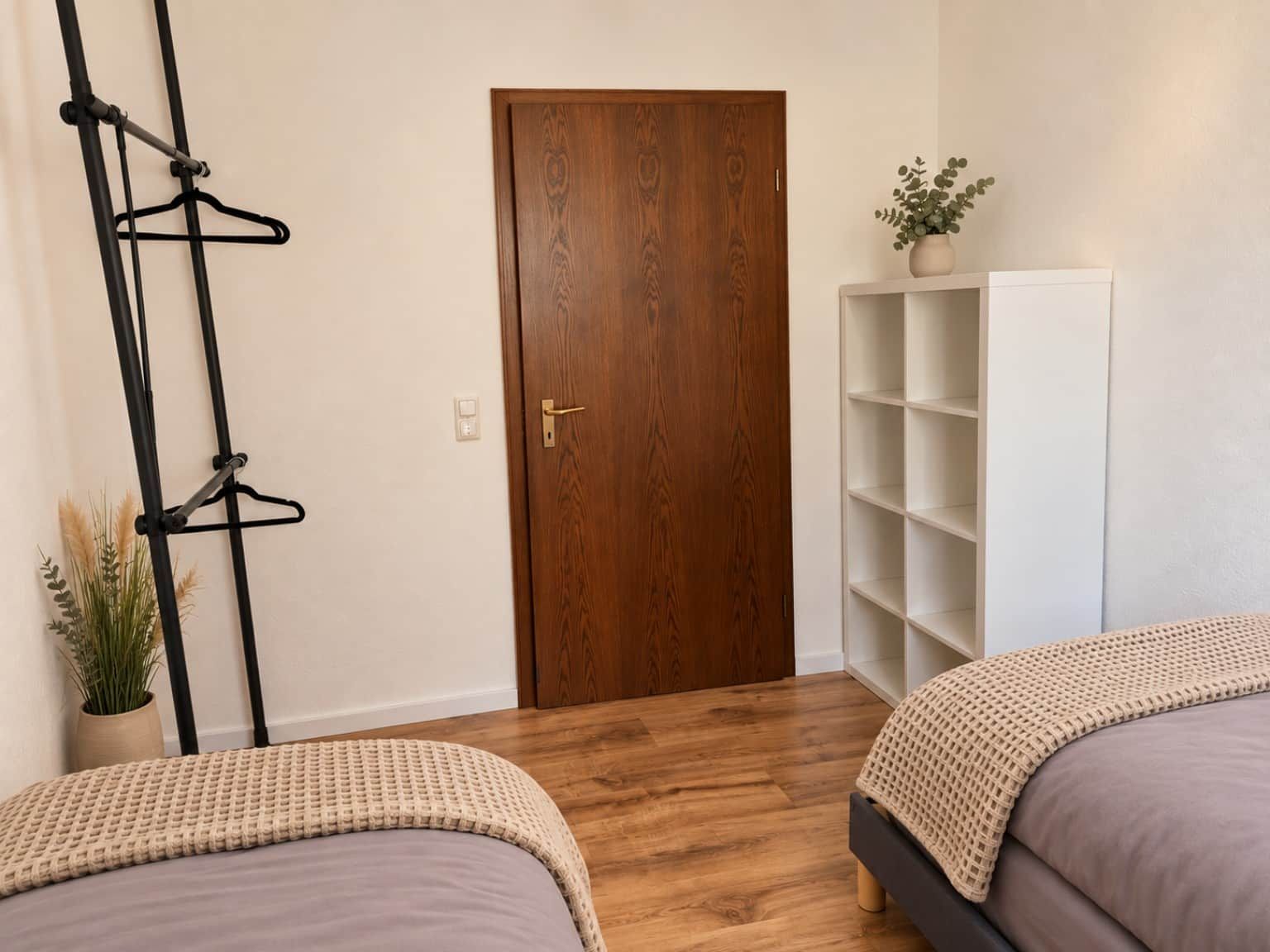 A room with two beds, a wooden door, a white shelving unit, and a black metal rack.