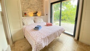 Una camera da letto con un letto, un comodino, una mensola a parete con un vaso e fiori, e un'ampia porta scorrevole in vetro che si apre su un giardino.
