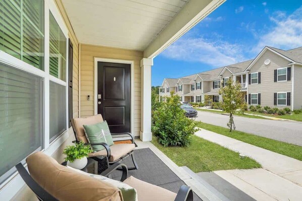 A porch with two chairs and a potted plant, a black door, and a view of a row of townhouses.