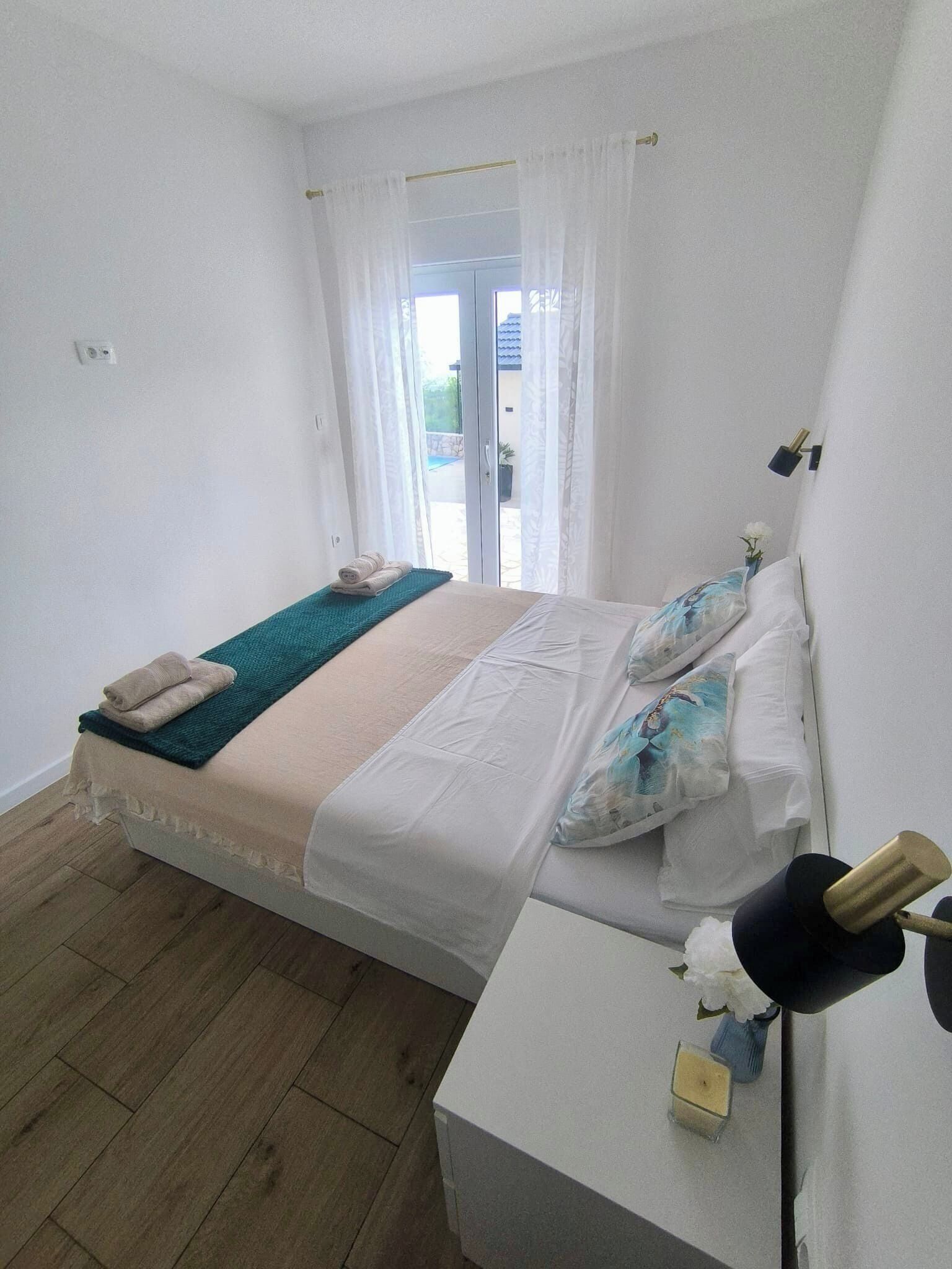 A bedroom with a bed, a bedside table, a lamp, and a view of the outside.