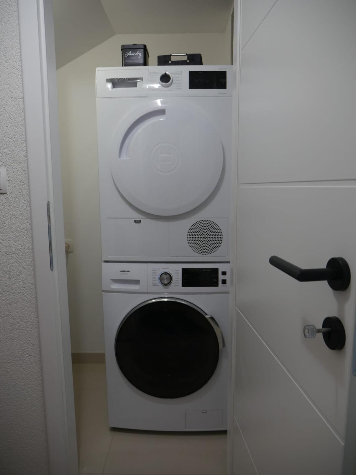 Ein kleiner Waschraum mit einer Waschmaschine und einem Trockner, einer kleinen Ablagefläche mit einem Wäschekorb und einer Tür mit Griff.