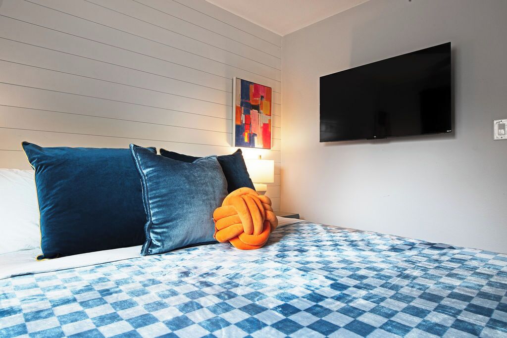 A hotel room with a checkered bedspread, a flat-screen TV mounted on the wall, and a colorful abstract painting above the bed.