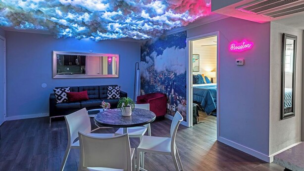 Een moderne woonkamer met een plafond dat lijkt op een wolk, een neonbord met daarop 'Houston' en een gezellige zithoek met een bank en stoelen.