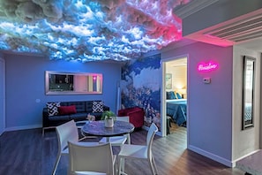 Een moderne woonkamer met een plafond dat lijkt op een wolk, een neonbord met daarop 'Houston' en een gezellige zithoek met een bank en stoelen.