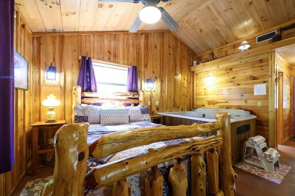 A wooden cabin interior with a bed, a window with curtains, a wooden headboard, a small table with a lamp, and a bathroom with a shower.