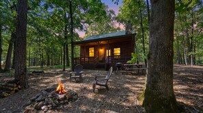 A cabin with a fire pit, two chairs, and a table in a forest setting.