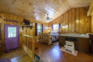 Interior de una cabaña con cama, bañera de hidromasaje, televisor y puerta de salida.