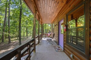 Un porche cubierto con barandillas de madera, una zona de estar con sillas y vistas a un área boscosa.
