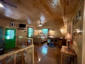 Interior de una cabaña con suelo de madera, una cama, un escritorio, una silla, un televisor y una puerta verde.