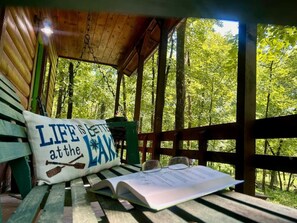 A wooden bench with a pillow and a book, set in a covered outdoor area with a view of trees.