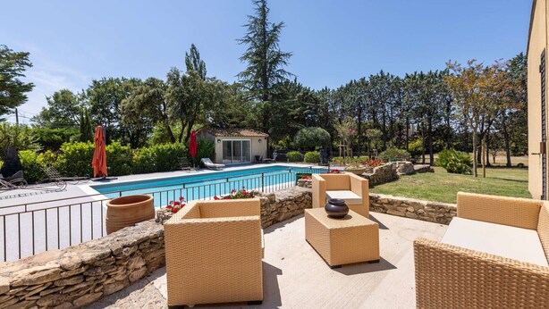 Un espace aménagé au bord de la piscine, avec des meubles en osier, un mur en pierre et une vue sur la piscine et les arbres environnants.