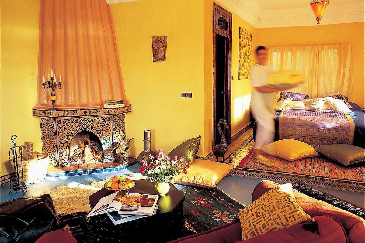 A room with a traditional fireplace, a bed with patterned bedding, and a person walking through the doorway.