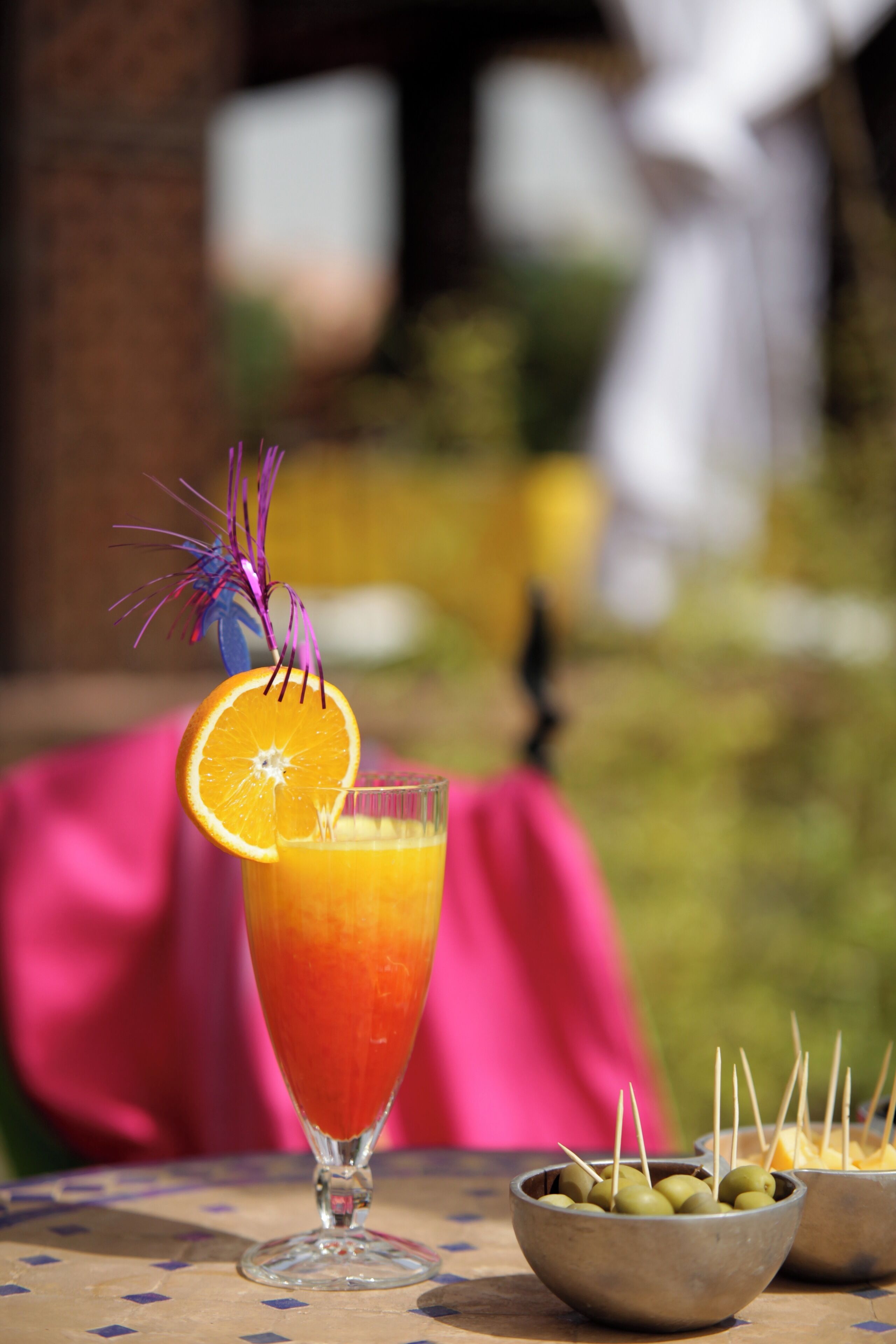 A glass of orange juice with a slice of orange and a decorative purple flower on a table.