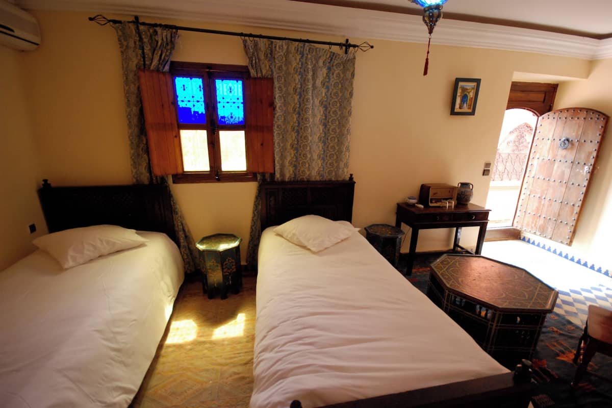 A room with two beds, a small table, a decorative cabinet, and a window with patterned curtains.