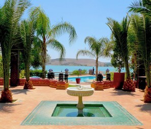 A pool area with a fountain, palm trees, and a view of a body of water. - RIVE DU LAC (Lalla Takarkoust)