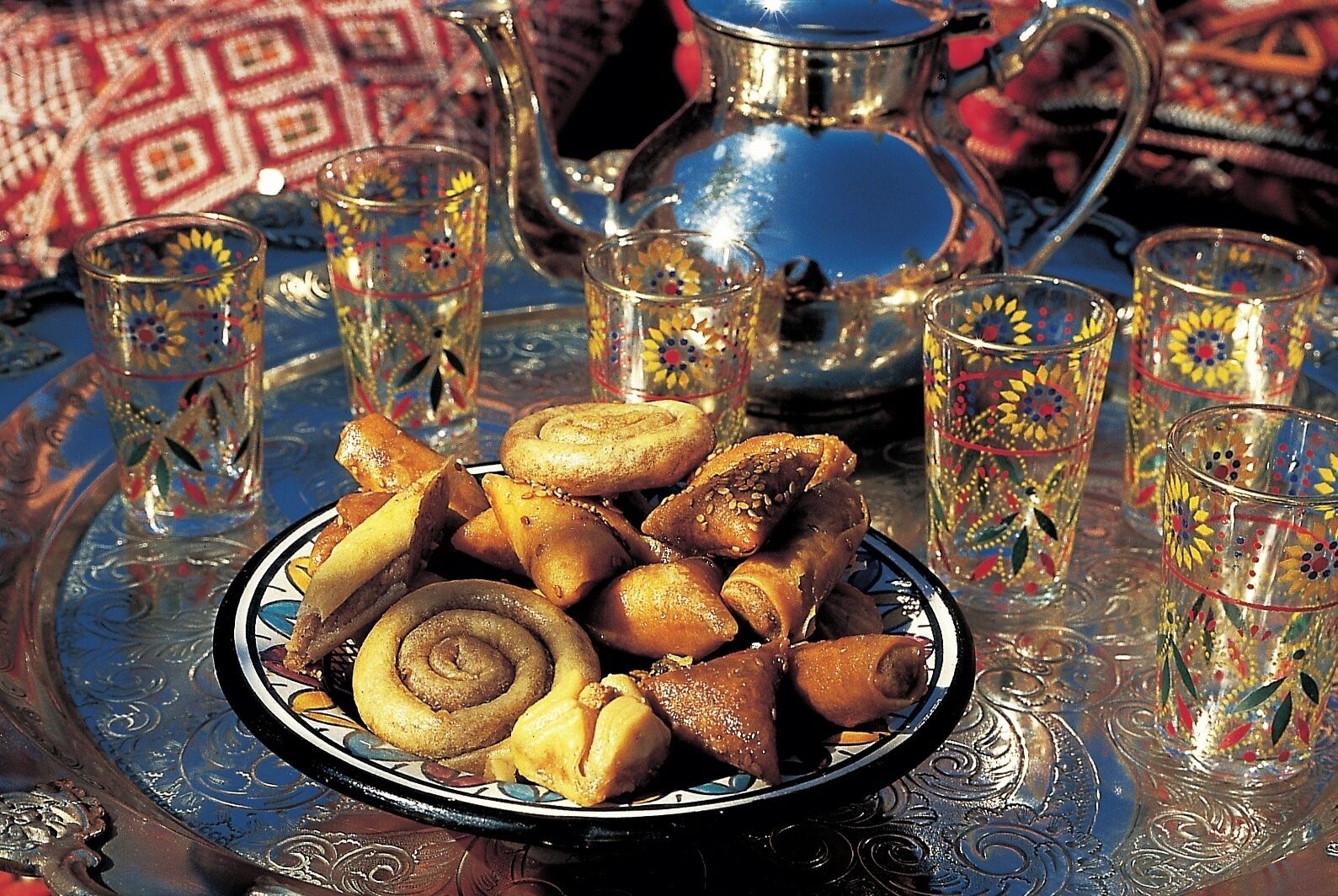 A tray with pastries and glasses of tea, set on a patterned tablecloth.