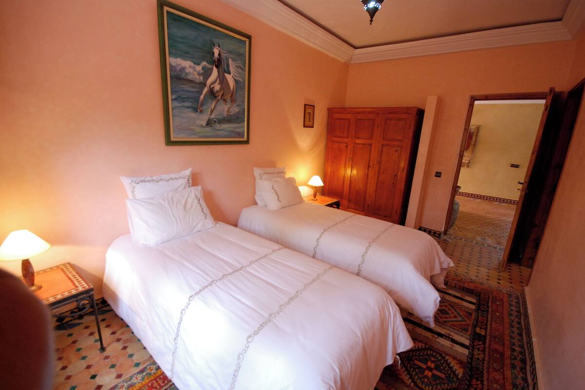 A hotel room with two beds, a painting on the wall, a wooden wardrobe, and a patterned carpet.