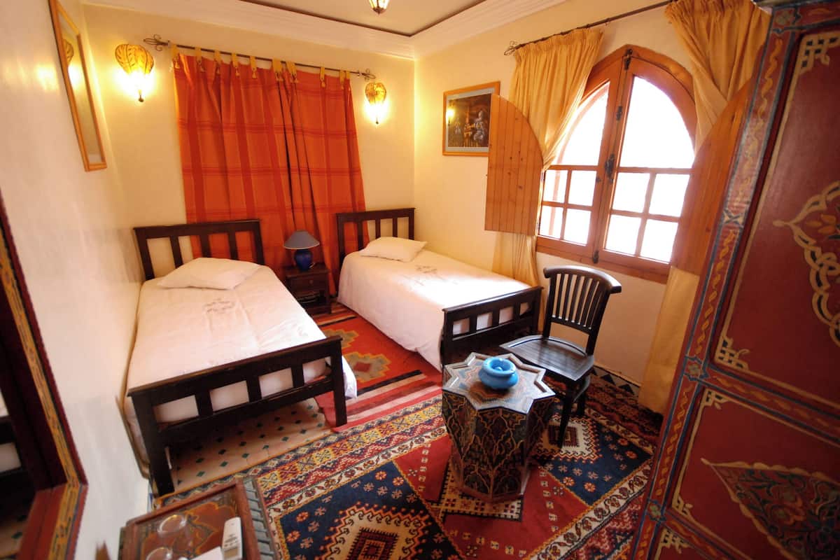 A room with two beds, a small table, a chair, and a window with curtains.