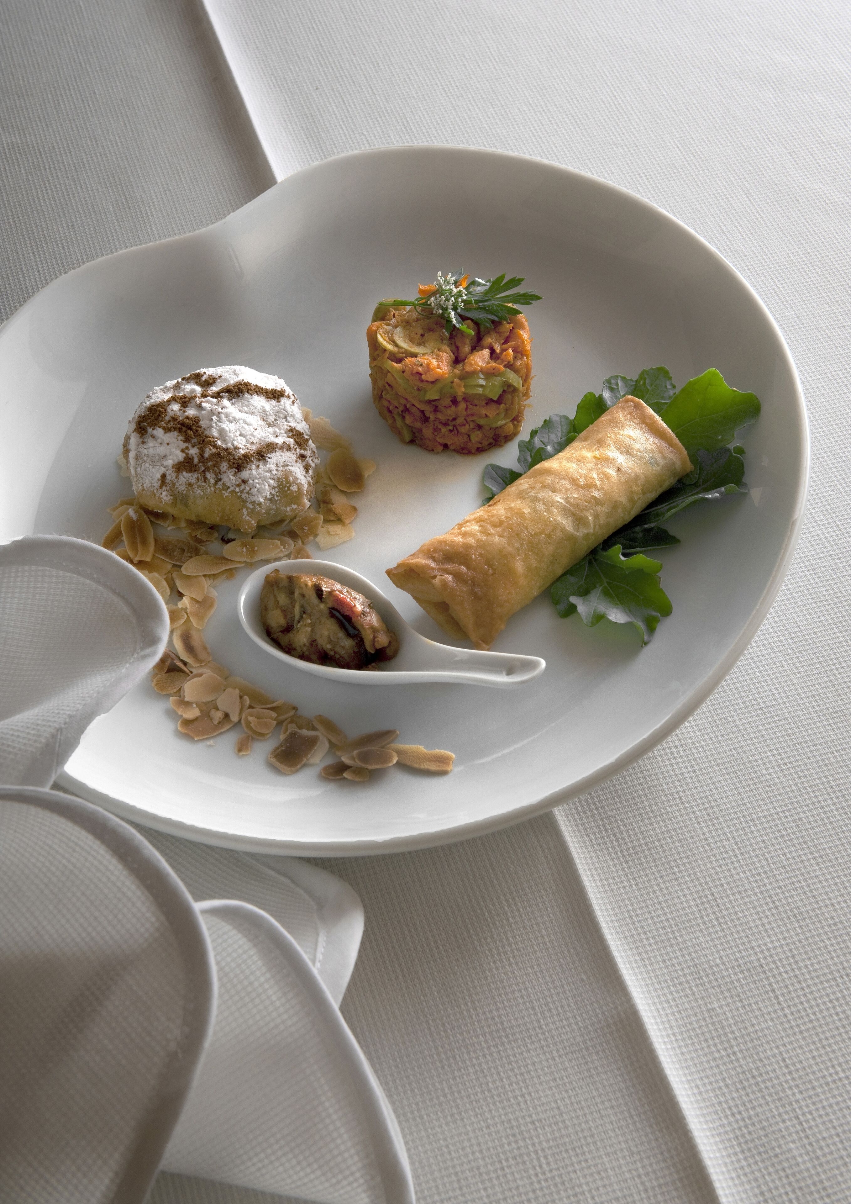 En vit tallrik med en dessert, en rullad bakelse och en liten skål, allt placerat på ett vitt bordsduksklätt bord.
