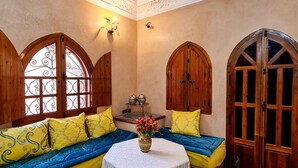 Une pièce avec une table ronde, des bancs en bois, un canapé avec des coussins jaunes et bleus, et un tapis à motifs.