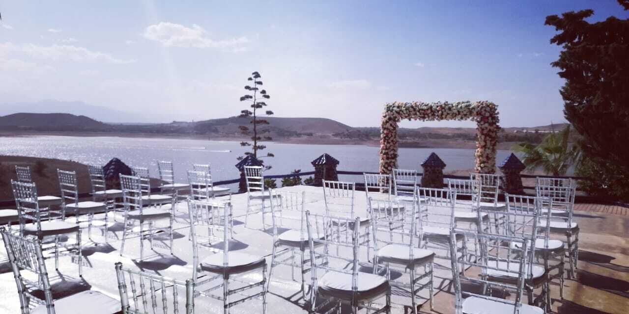 An outdoor seating area with white chairs and tables, a decorative arch, and a view of a body of water and hills in the background.