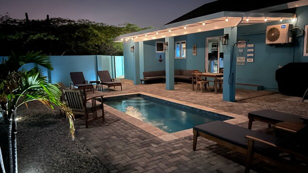 A pool area with lounge chairs and a table, surrounded by a blue wall and a covered patio.