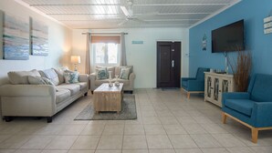 A living room with a beige sofa, a blue armchair, a wooden coffee table, and a TV on the wall.