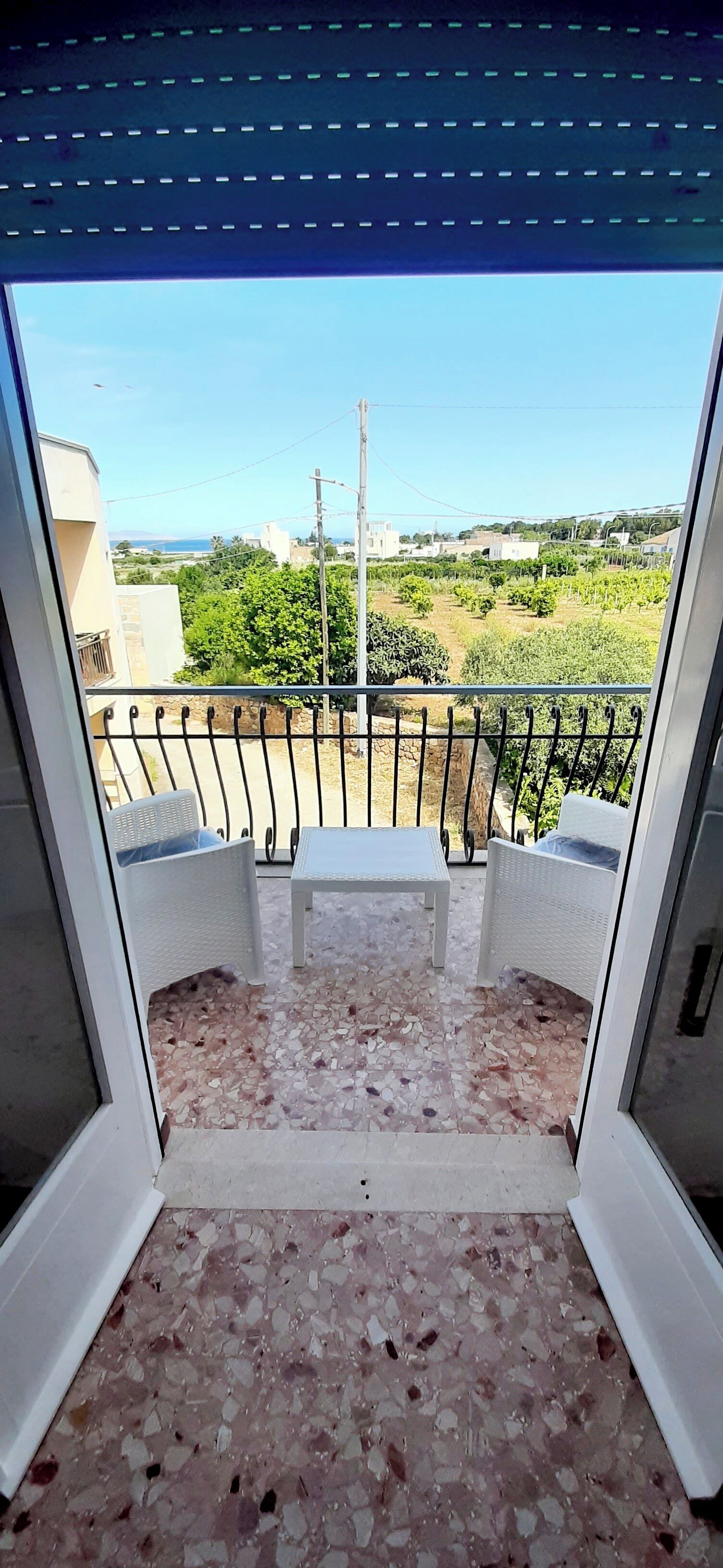 A balcony with a terrazzo floor, two white chairs, and a view of a vineyard and buildings in the distance.