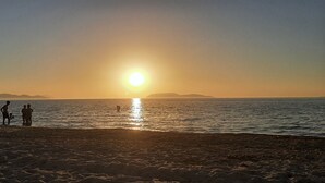 A beach scene with the sun setting over the water, people standing and walking along the shore, and a beach umbrella providing shade. - The house of the islands Sea View ( The House Of The Islands ) (Marausa Lido)