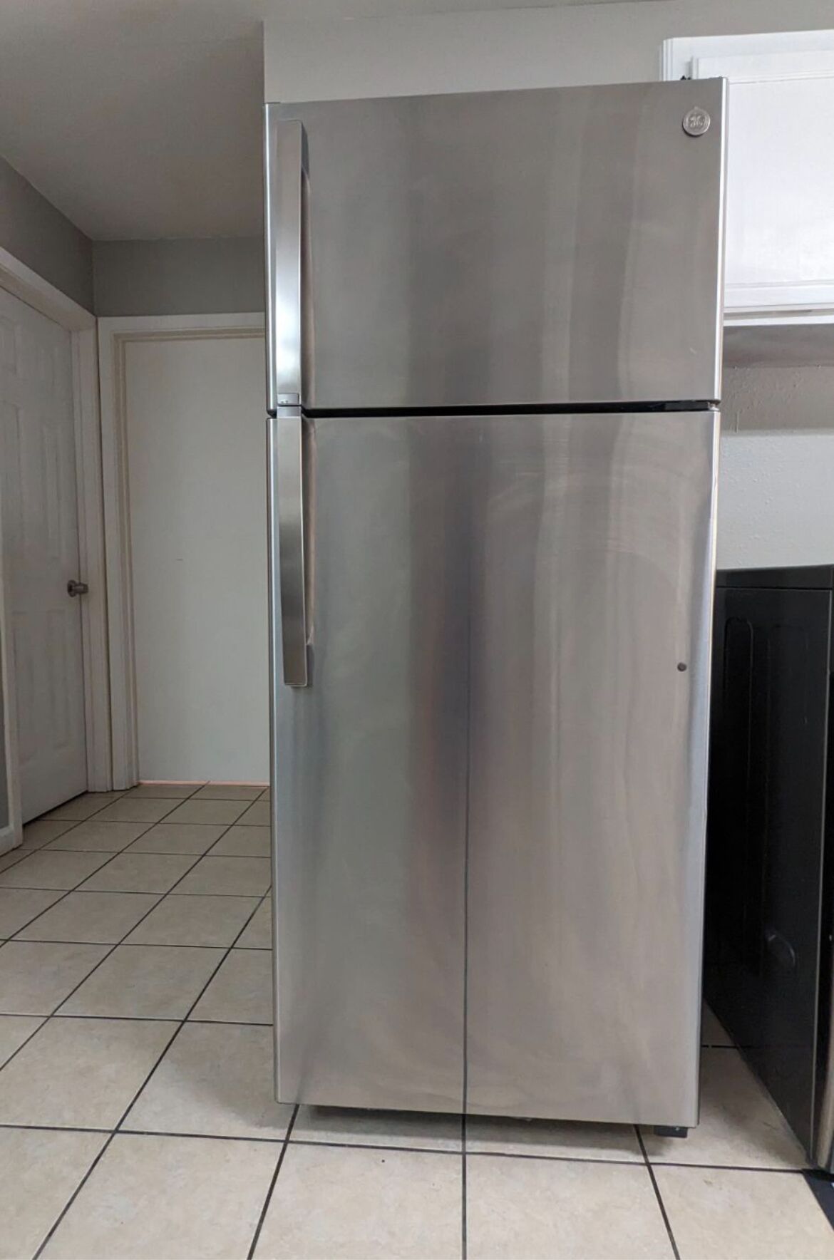 A stainless steel refrigerator in a kitchen with tiled flooring and a white door in the background.
