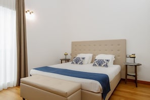 A bedroom with a large bed, two bedside tables with lamps, a window with curtains, and a wooden floor.