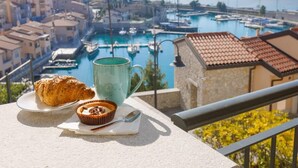 A balcony with a croissant, muffin, and mug, overlooking a marina with boats and a coastal town.
