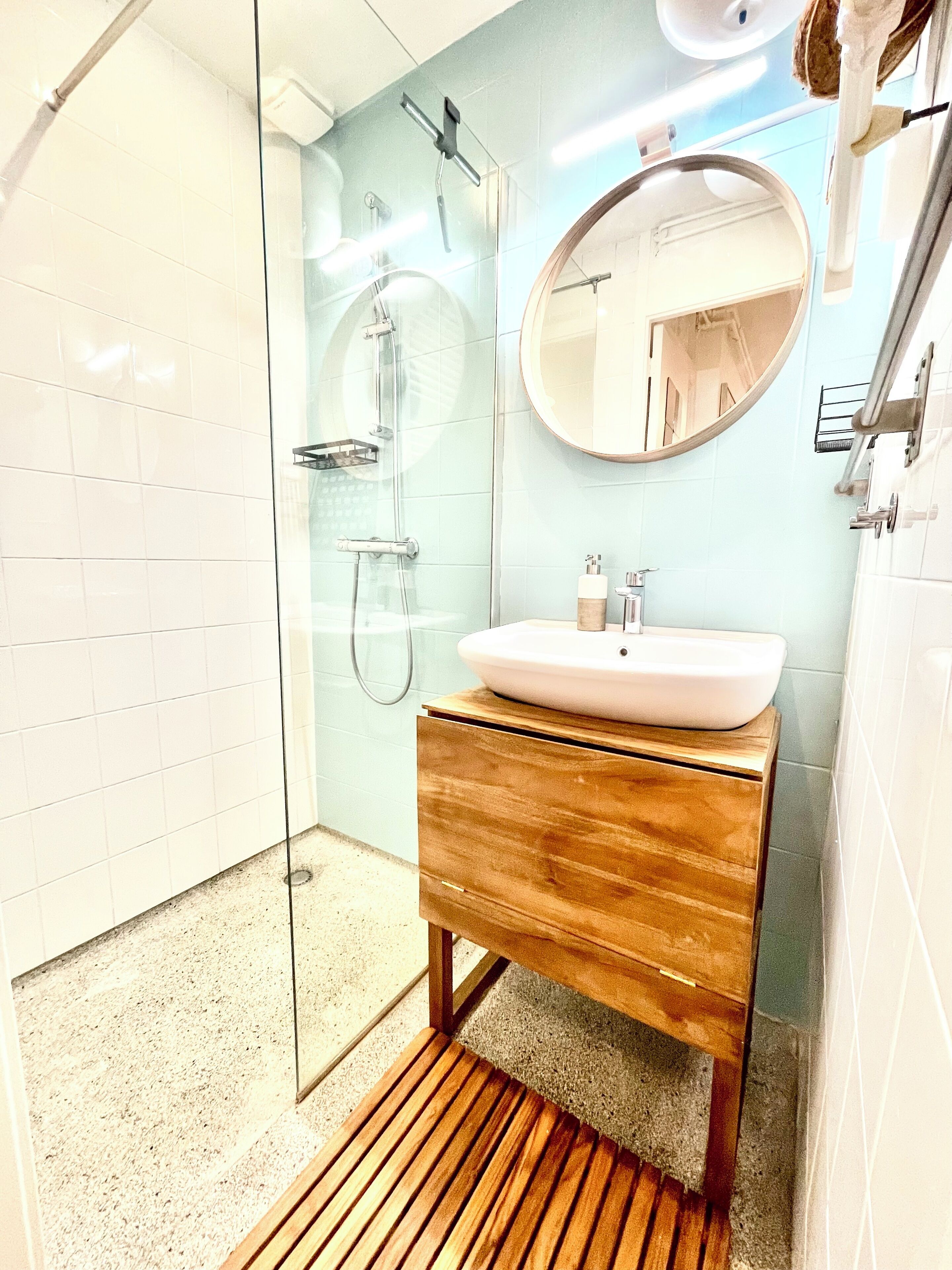 Un baño moderno con ducha de vidrio, tocador de madera con lavamanos blanco y un espejo redondo.