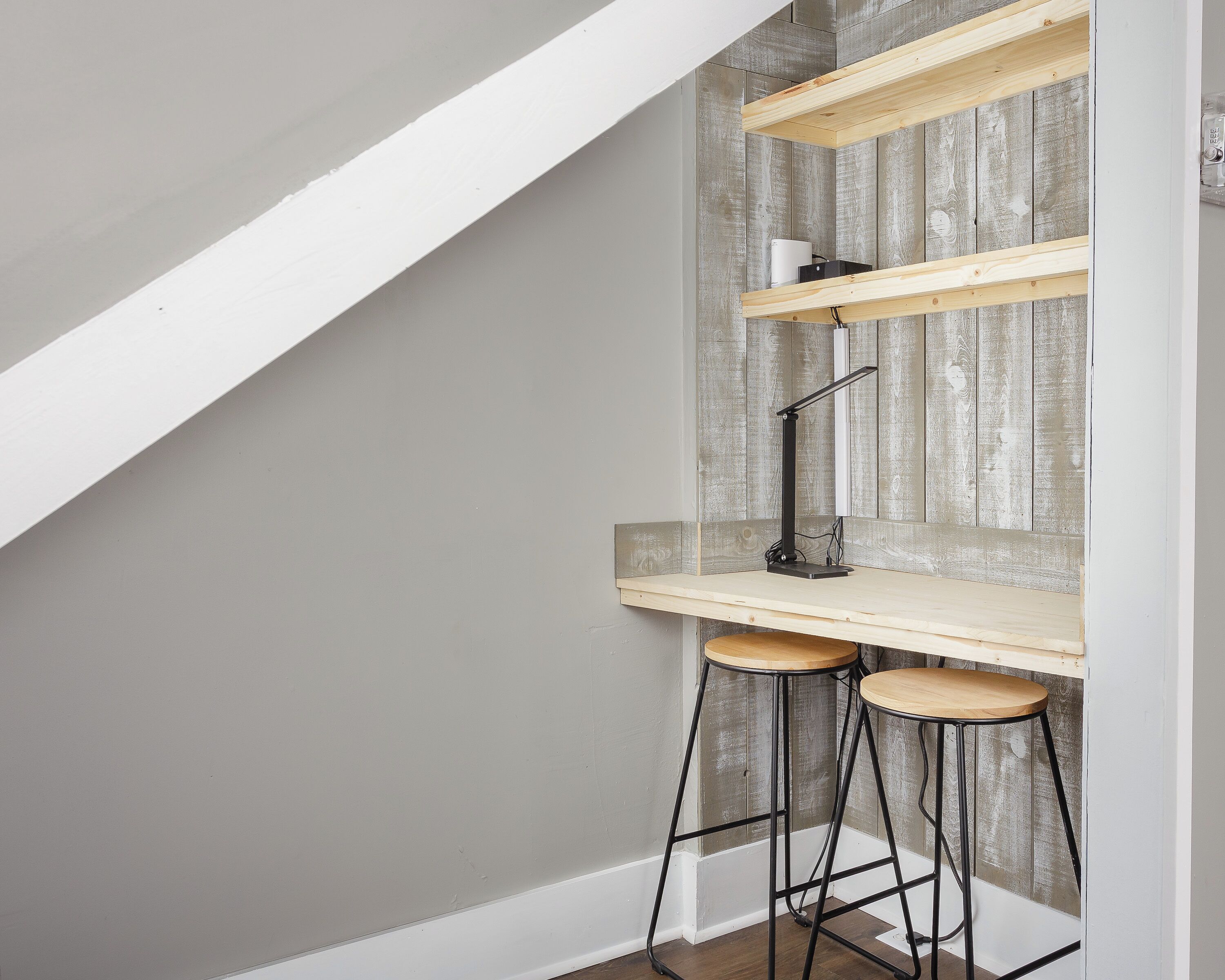 Un espace de travail aménagé dans un coin, avec un bureau, deux tabourets et une étagère le long d’un mur en bois.