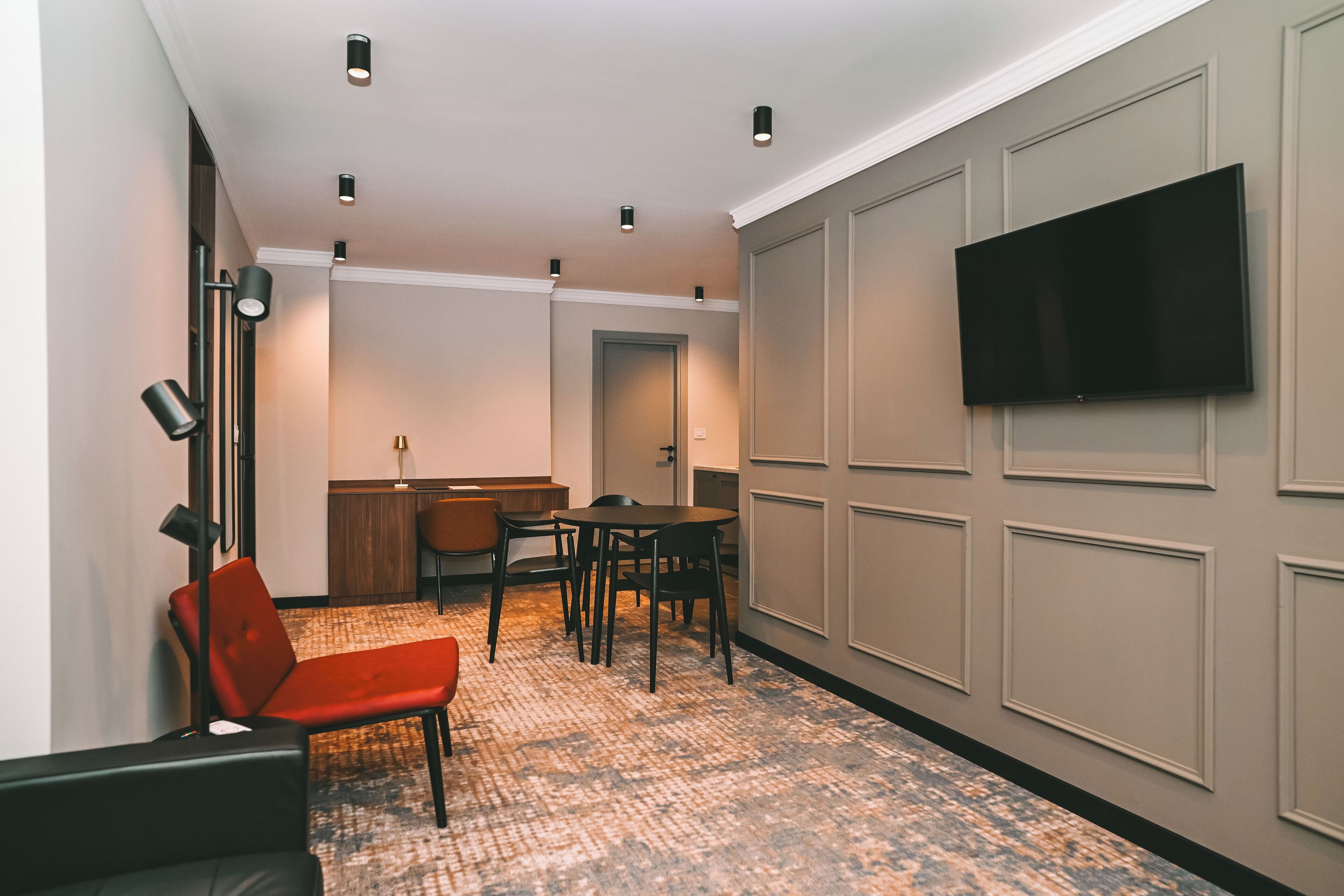 Una habitación de hotel con una televisión de pantalla plana montada en la pared, un rincón de comedor con una mesa y sillas, y una silla roja.