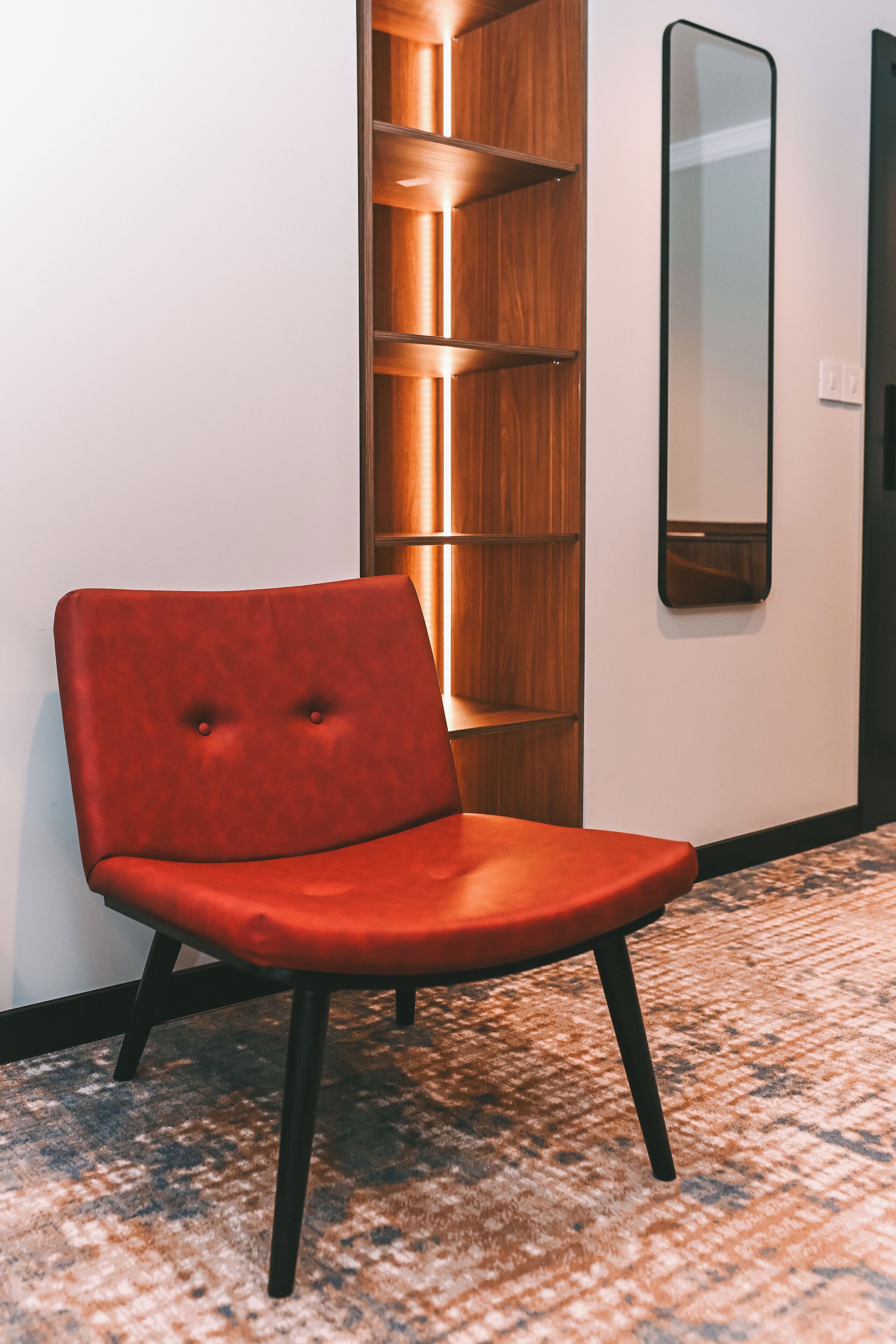 A red upholstered chair with black legs in a hallway with a patterned carpet, a wooden shelving unit, and a mirror on the wall.